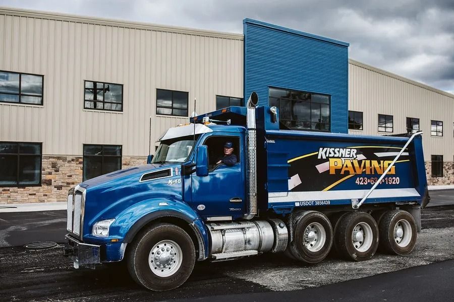 Blue Kissner Paving dump truck with driver, parked in front of a building with a blue accent wall.