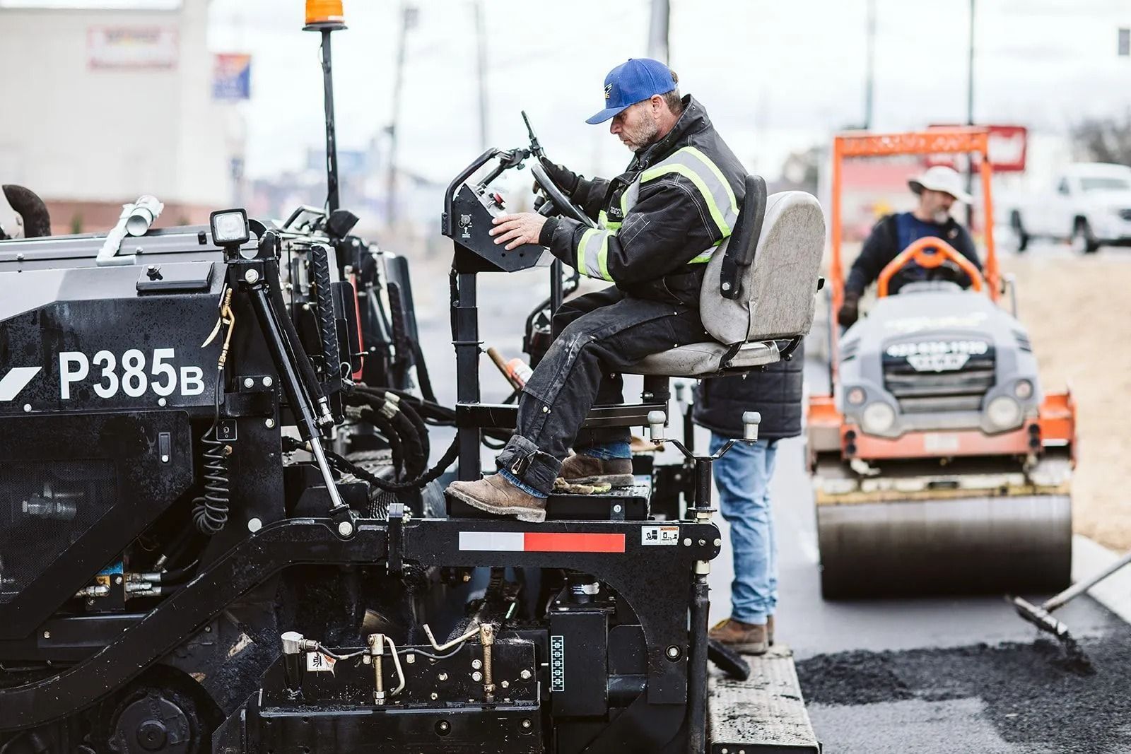 Man operating asphalt paving machine on a road; another worker in the background uses a roller.