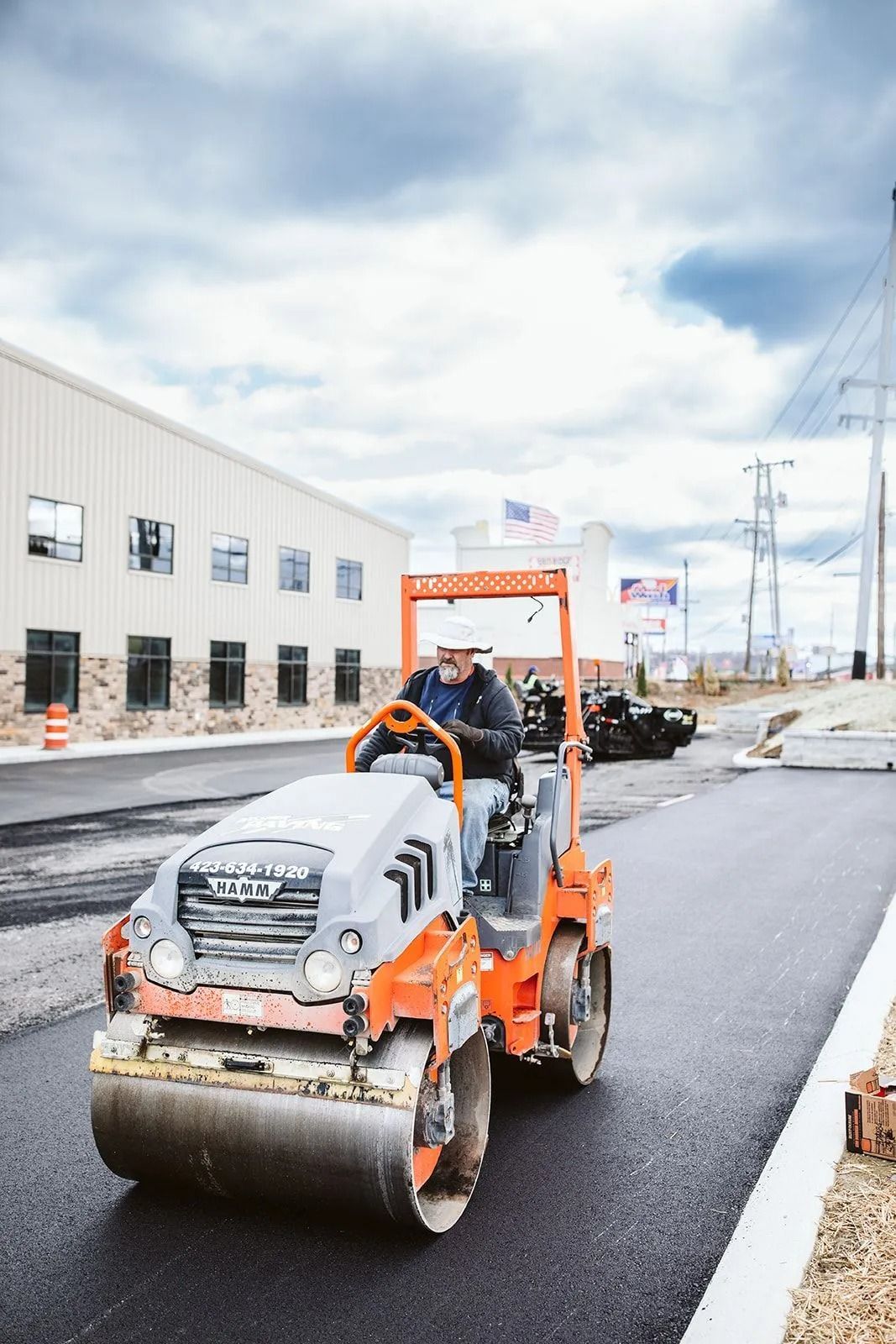 Man operating a Hamm roller to pave a road near a building and power lines.