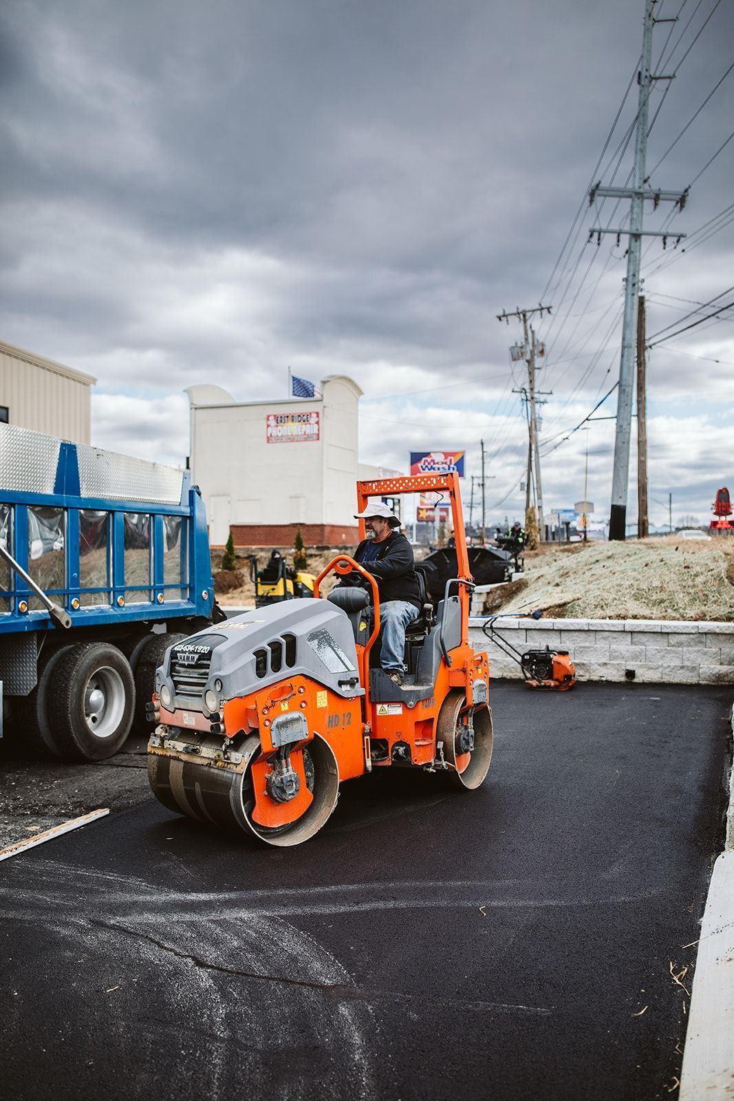 Worker operating a compact asphalt roller on a newly paved parking lot. Orange equipment, cloudy sky.