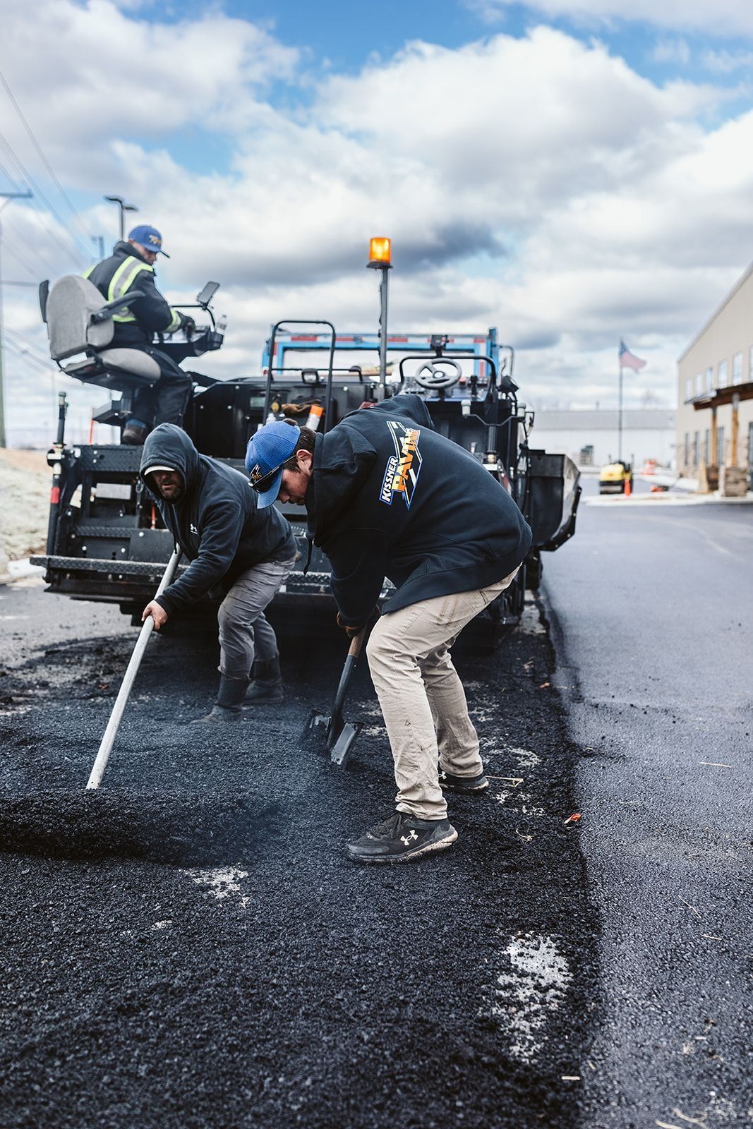 Road workers laying asphalt. One operates the machine, two manually spread the asphalt on a sunny day.
