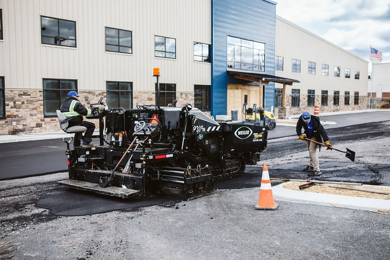 Asphalt paving machine laying asphalt on a road with a worker using a shovel, in front of a building.