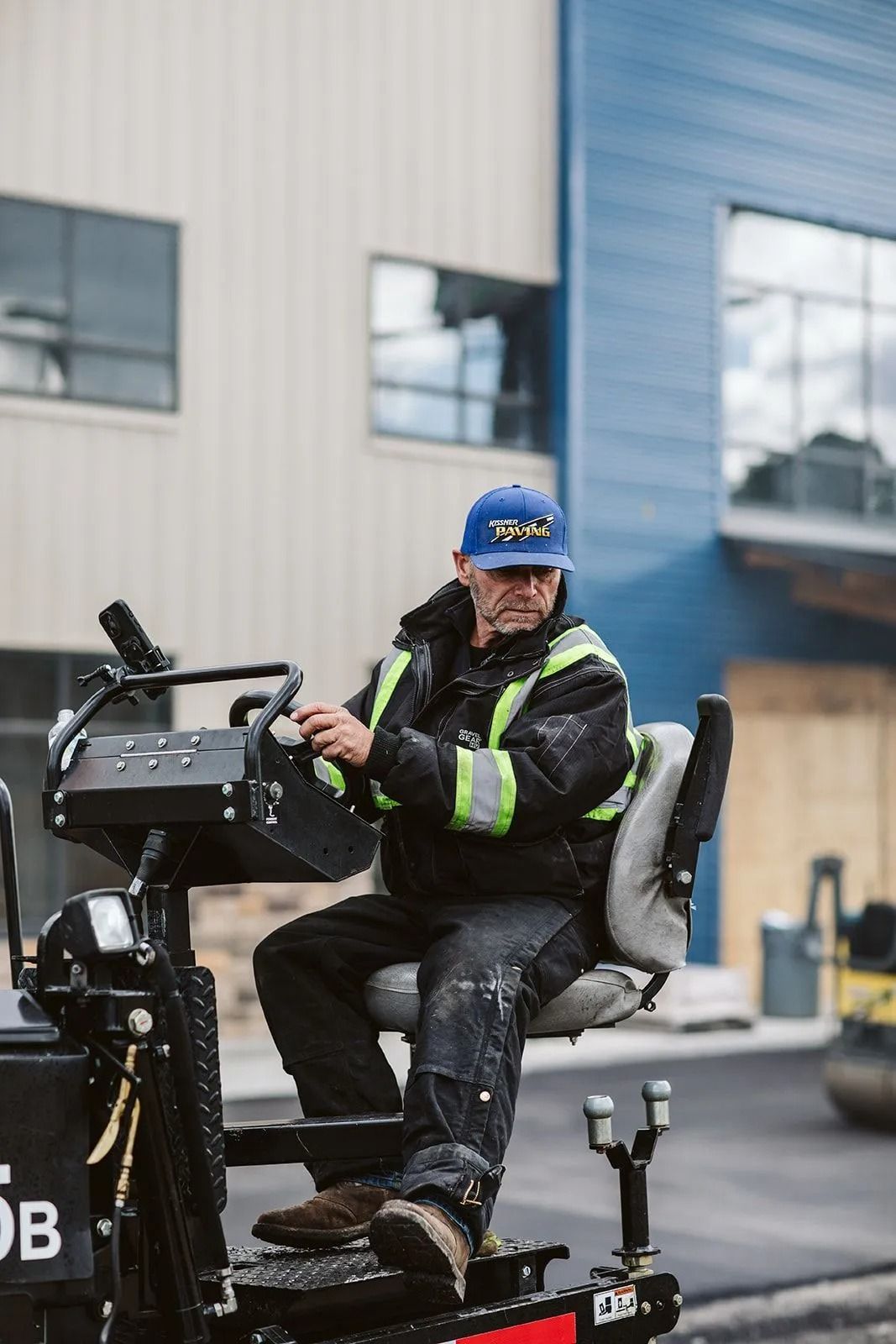 Man operating paving machine. He wears a cap, jacket with reflective stripes, and is focused on the task.