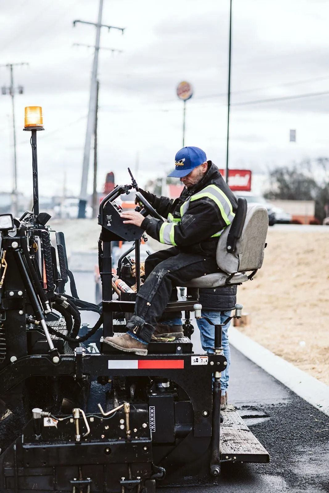 Man operating asphalt paving machine on a road, wearing blue hat and reflective vest.