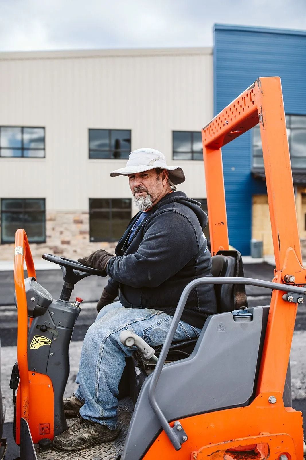 Construction worker operating a compact roller, wearing a white hat and gloves. Building in the background.