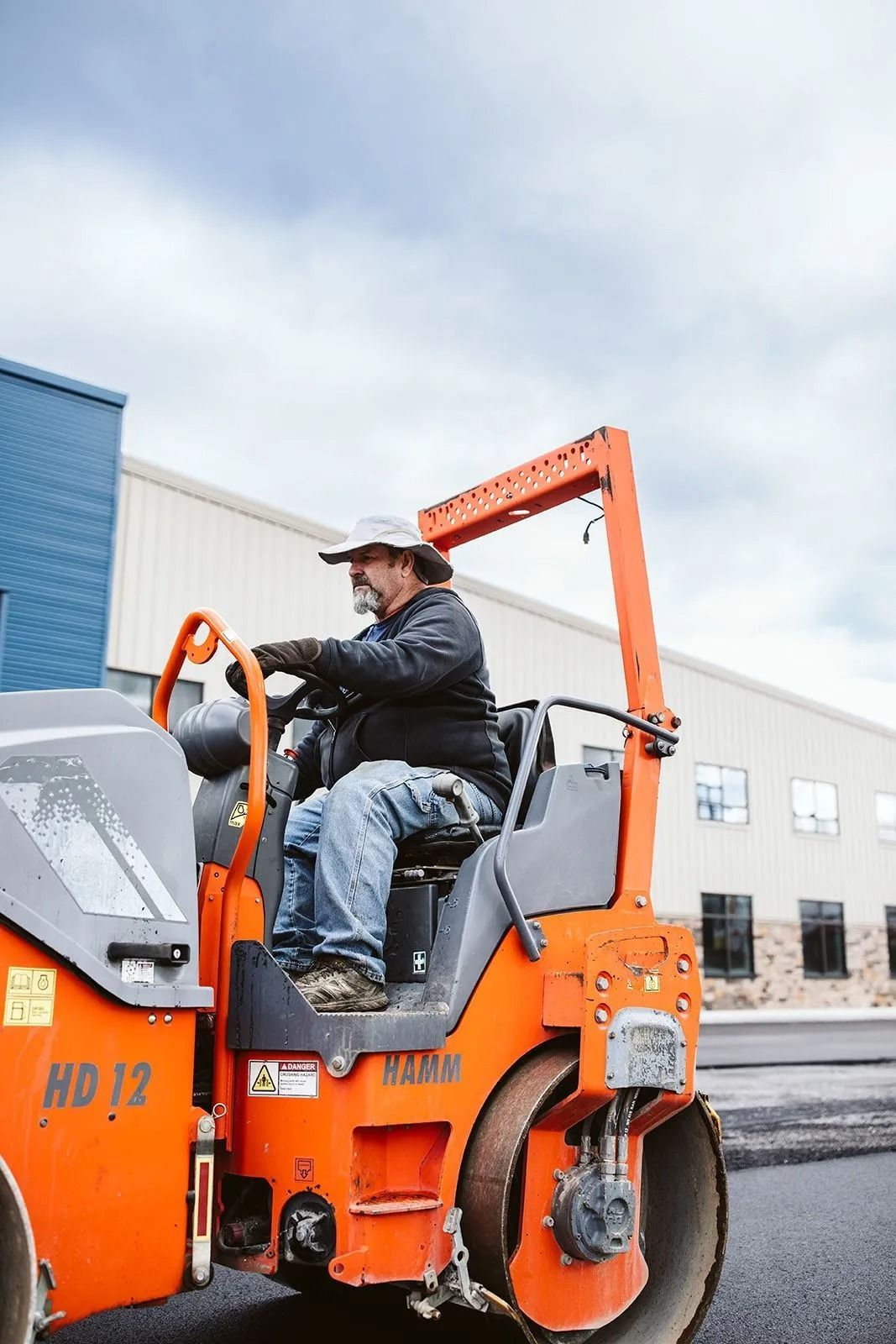 Man operating an orange road roller to pave asphalt. Cloudy day, factory building in background.