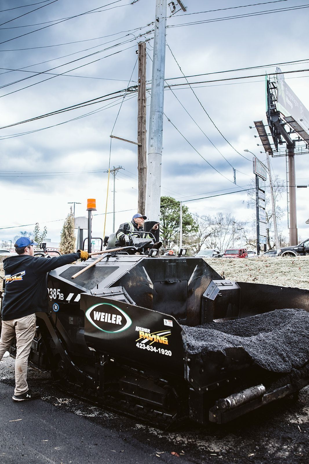Road paving crew working with asphalt paver near power lines and billboards.