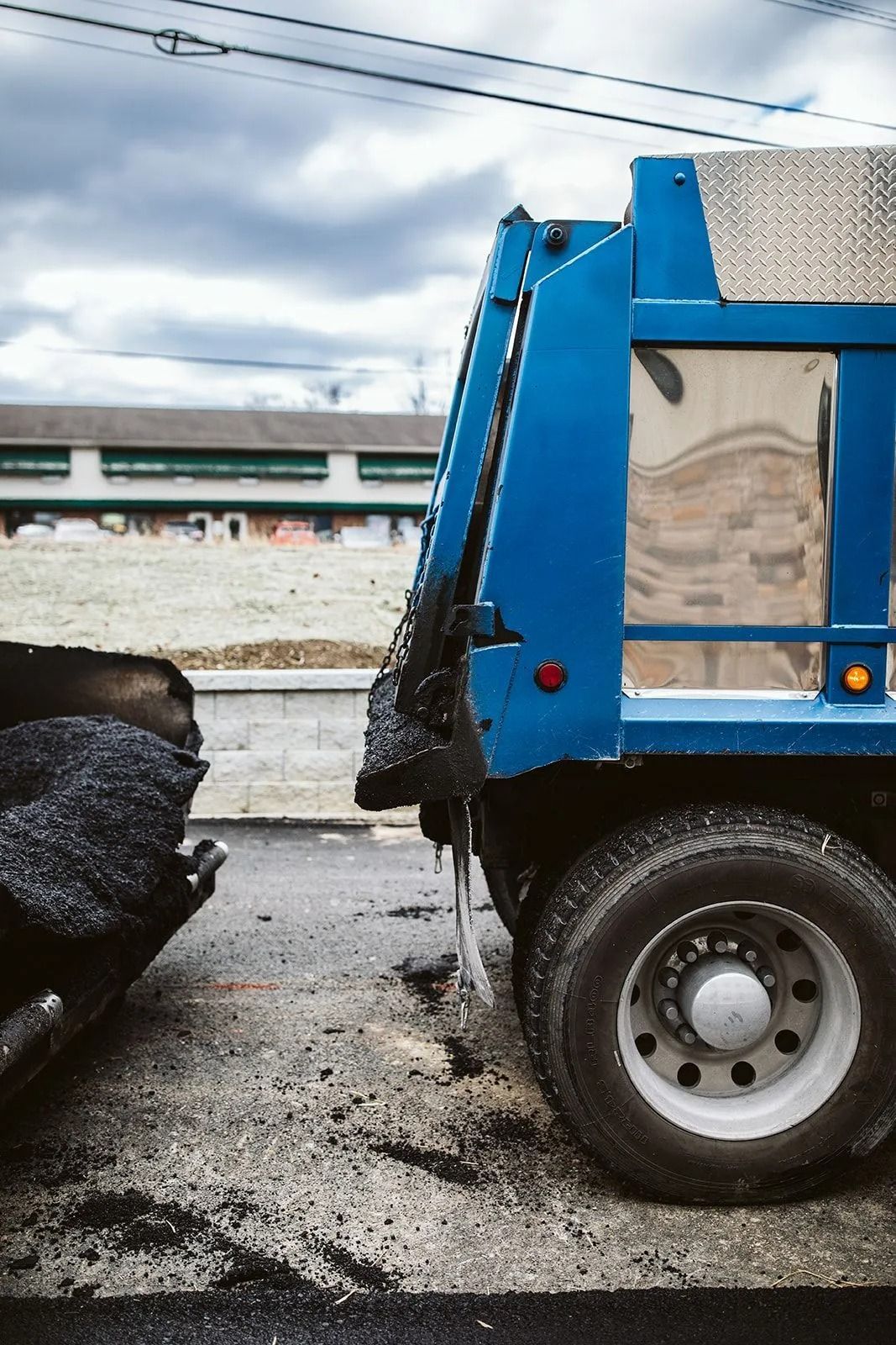 Blue dump truck dispensing asphalt onto a road under cloudy skies.