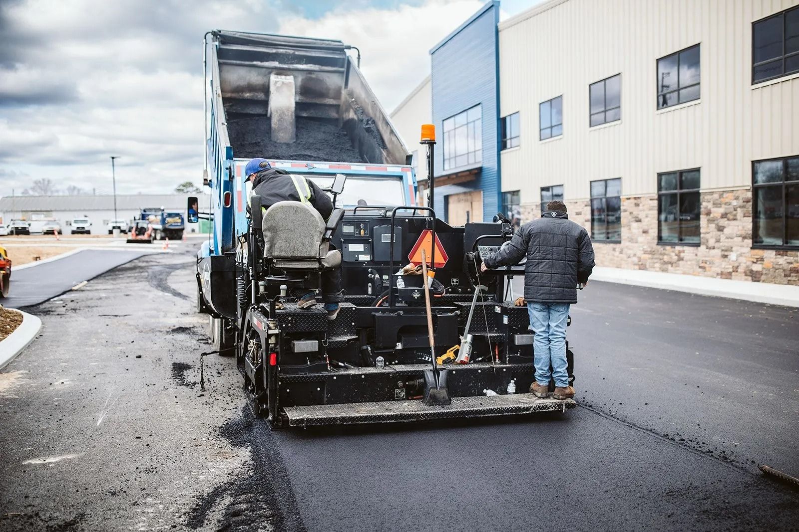 Asphalt paving: Workers operating machinery, pouring black asphalt on road. Blue dump truck, industrial building backdrop.