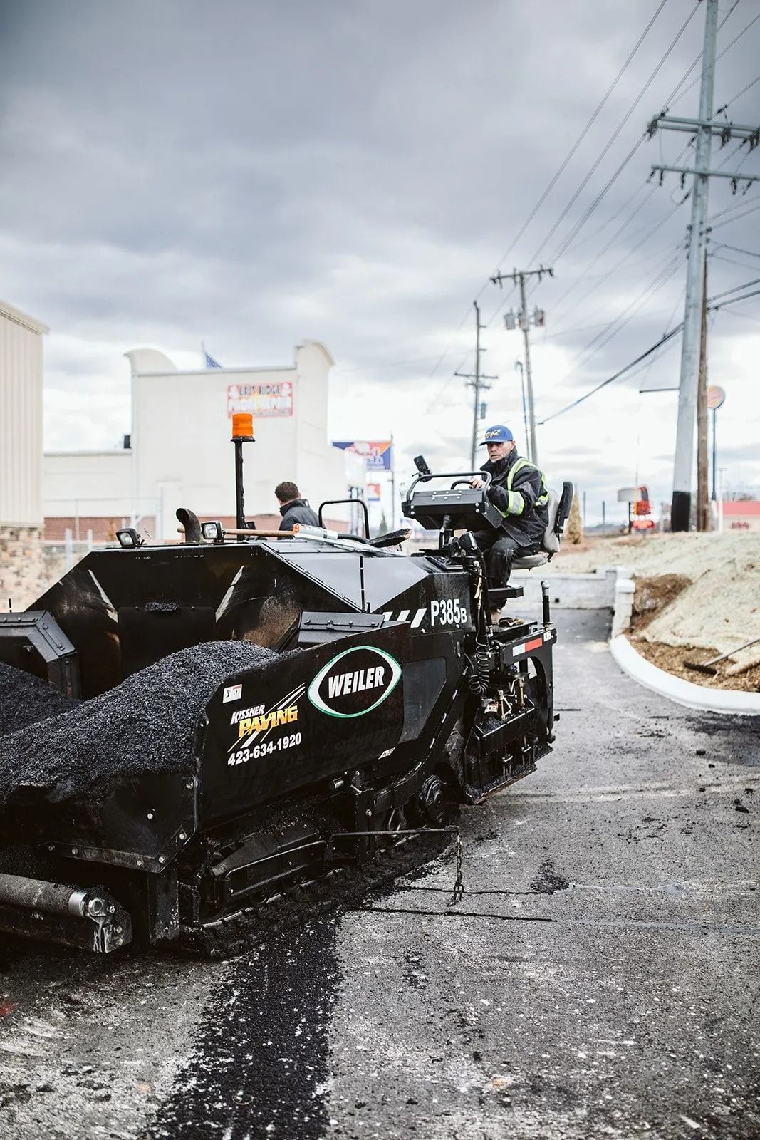Asphalt paving machine laying fresh black pavement. Worker operates on a street. Cloudy day.