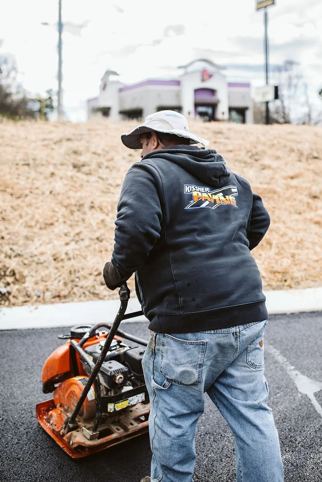Man compacting asphalt with a plate compactor outside, near a Taco Bell restaurant.
