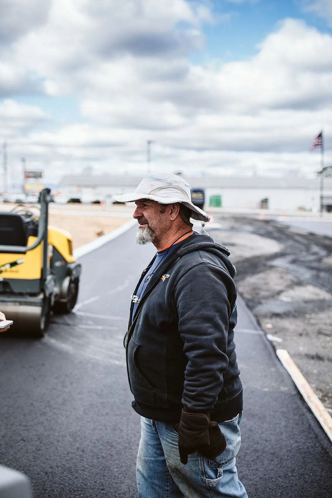 Man in hat, jacket, and gloves watches a steamroller smooth asphalt on a cloudy day.