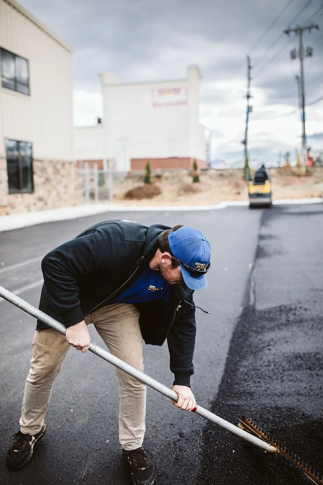 Man raking asphalt in front of a building under a cloudy sky, wearing a blue cap and jacket.