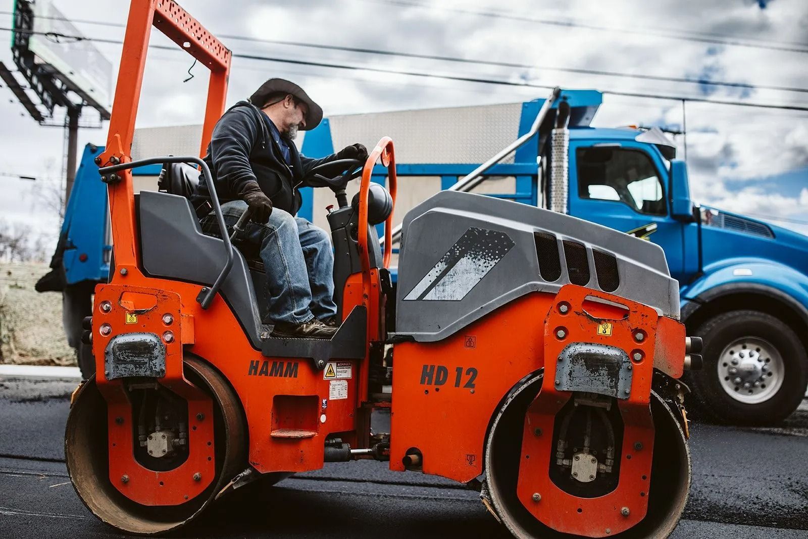 Man operating an orange road roller on asphalt with a blue truck nearby.
