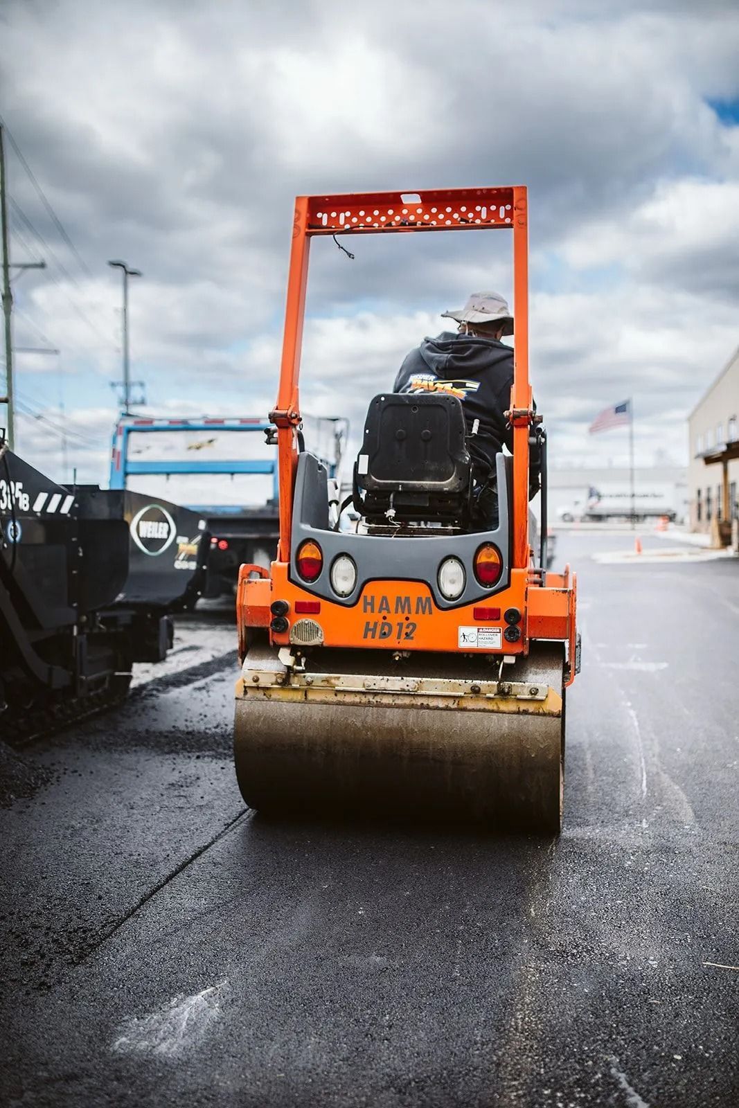 Orange road roller compacting asphalt, worker visible. Outdoors under cloudy sky.