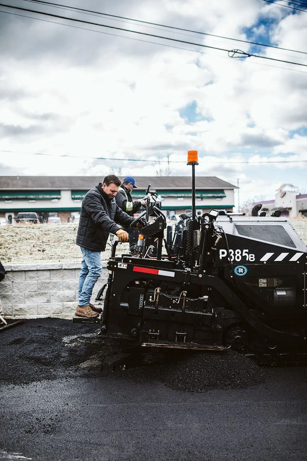 Two men operating a road paving machine on a wet, asphalt surface under a cloudy sky.