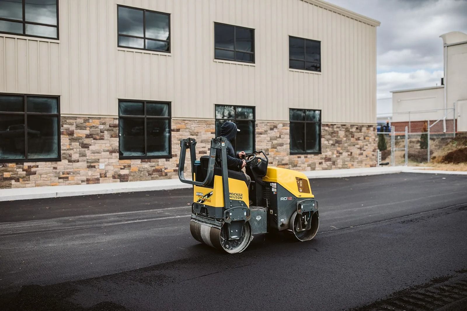 A small, yellow asphalt roller compacts a fresh black road in front of a building.