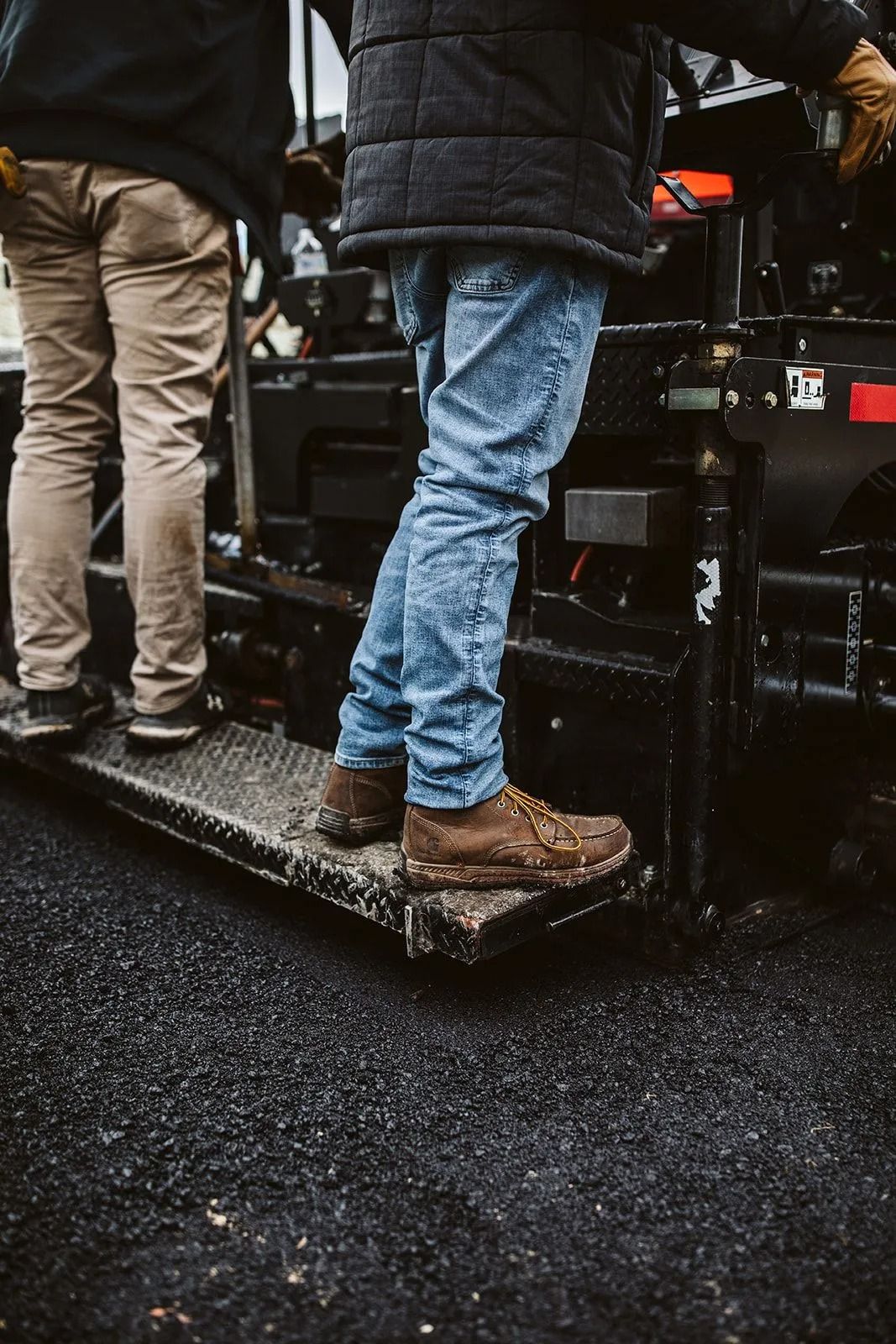 Two workers standing on an asphalt paving machine, one in blue jeans, the other in tan pants.