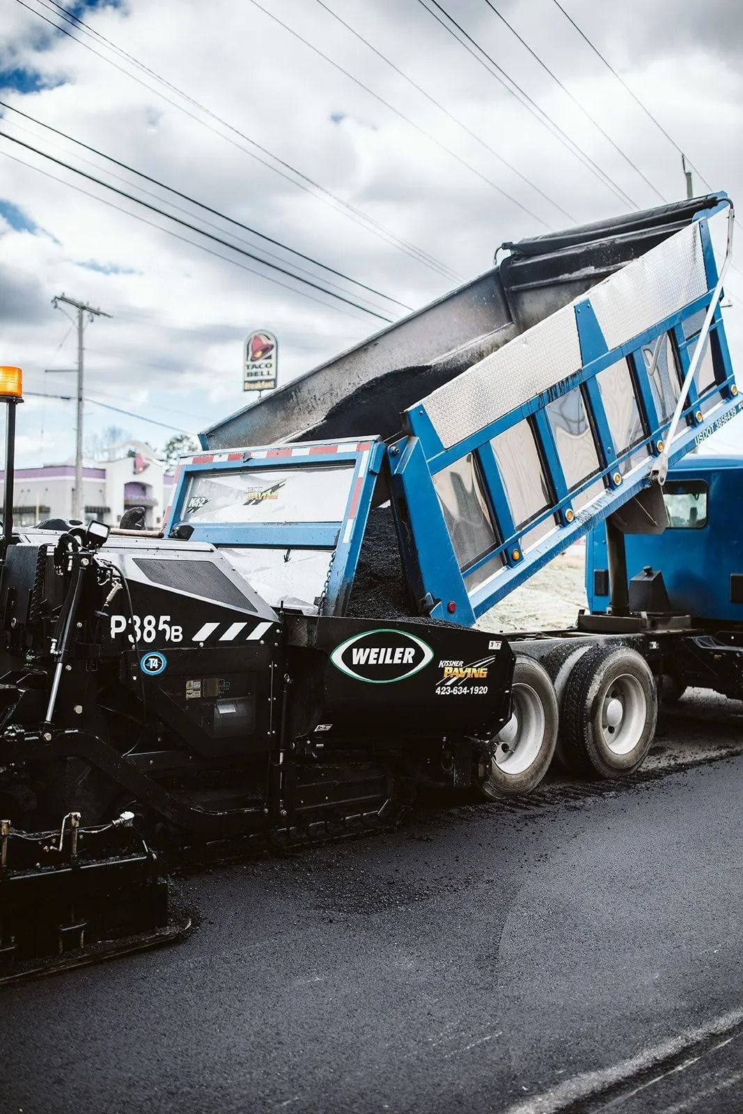 Truck dumping asphalt into a paving machine, on a road under a cloudy sky.