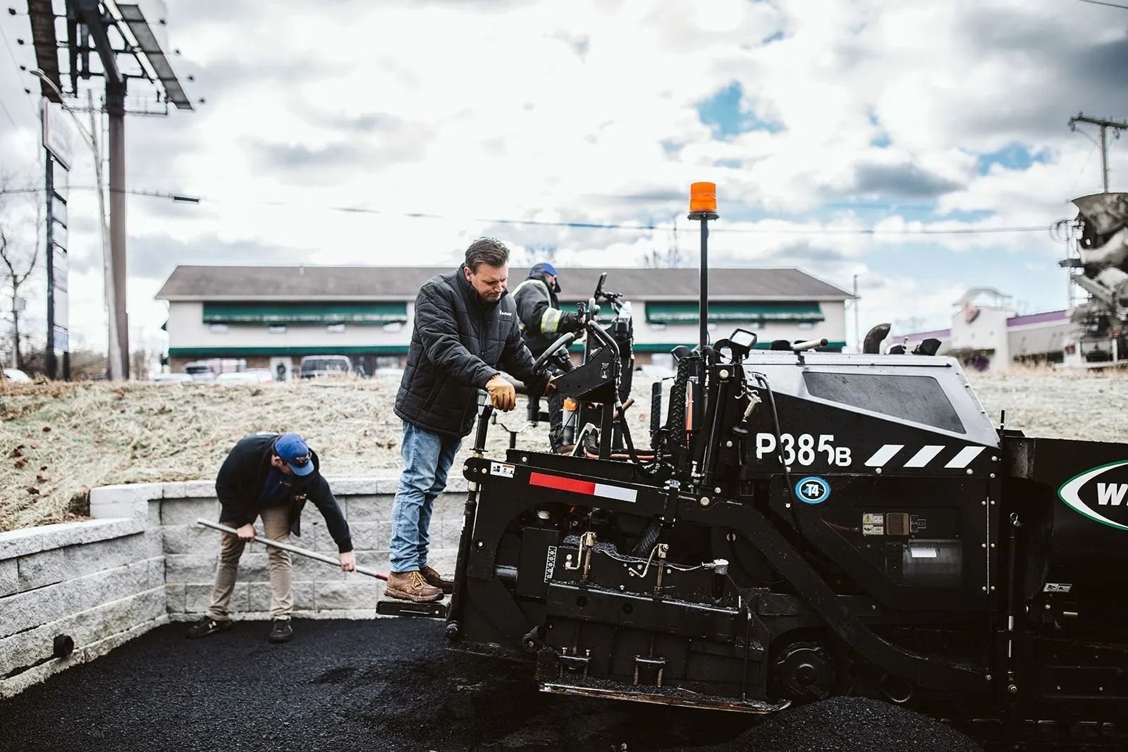 Two men paving with a machine on a cloudy day. One is using a tool, the other is operating machinery.