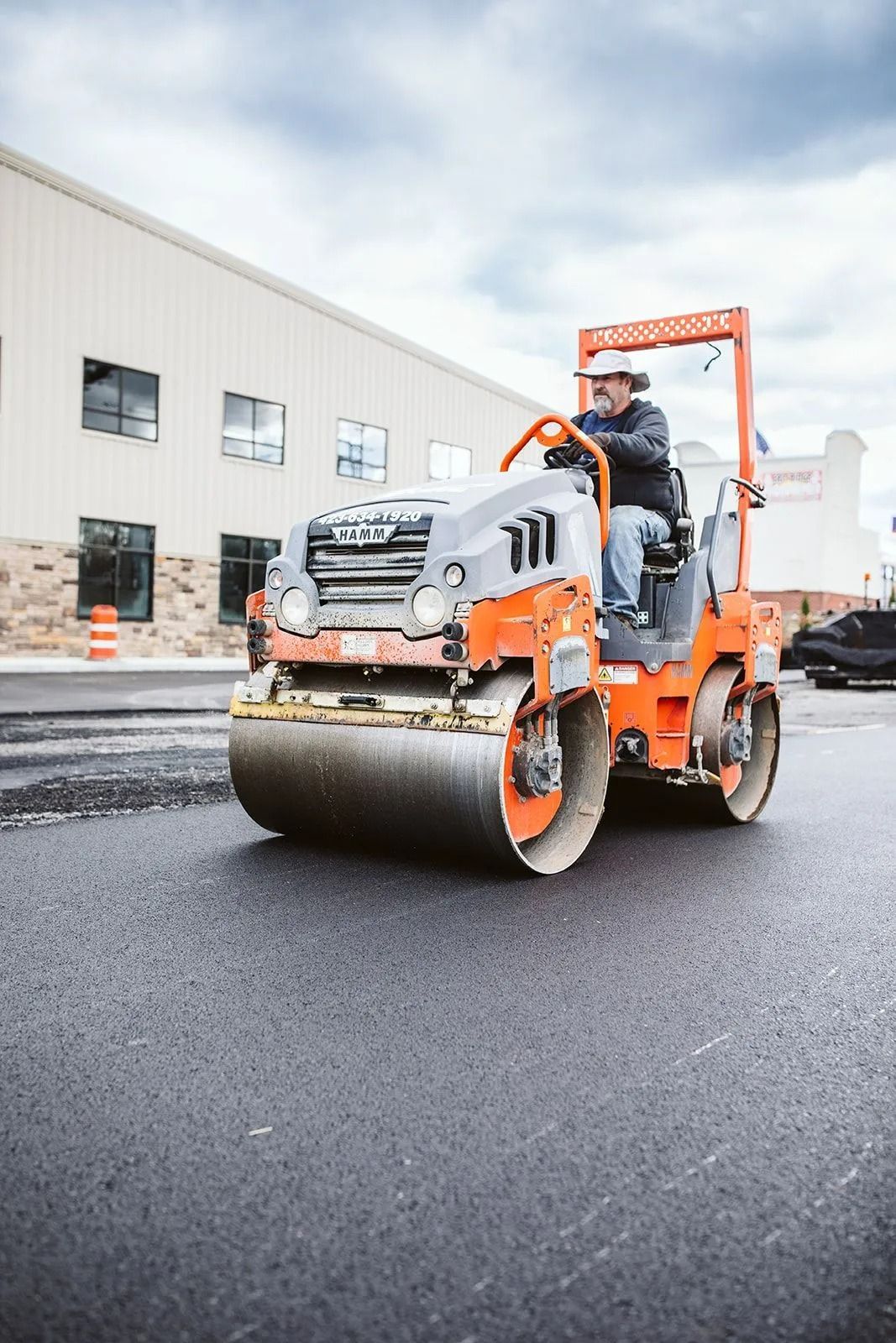 A construction worker operating an orange road roller on fresh asphalt in front of a commercial building.