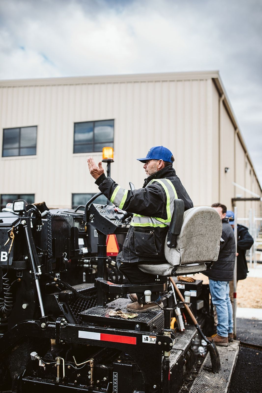 Man in a construction vehicle giving hand signals; a new road being built near a building.