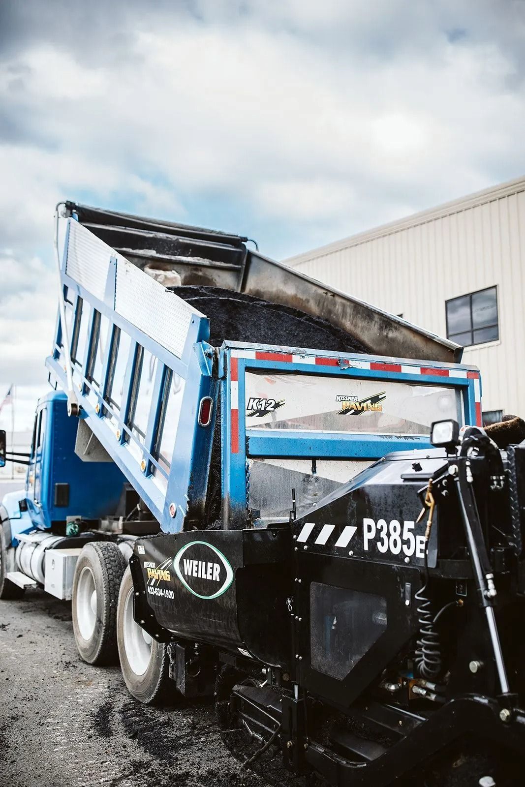Blue dump truck unloading asphalt into a road paving machine.
