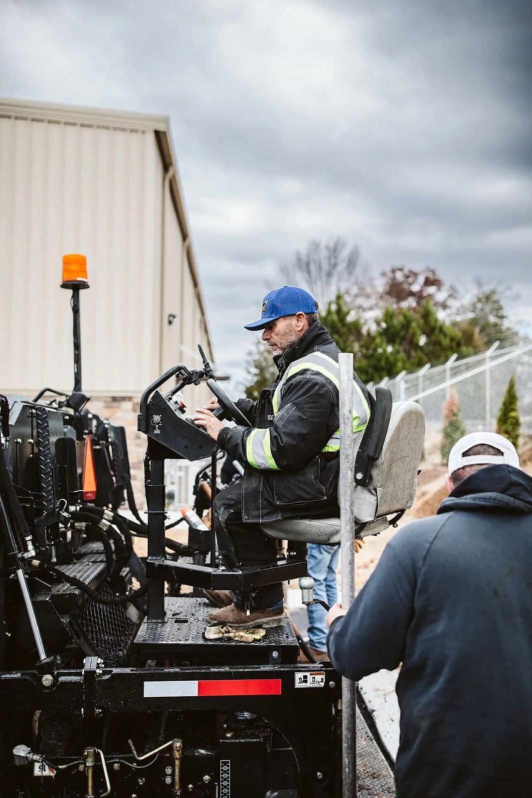 Man operating asphalt paving machine, another man assisting in industrial setting. Overcast sky.