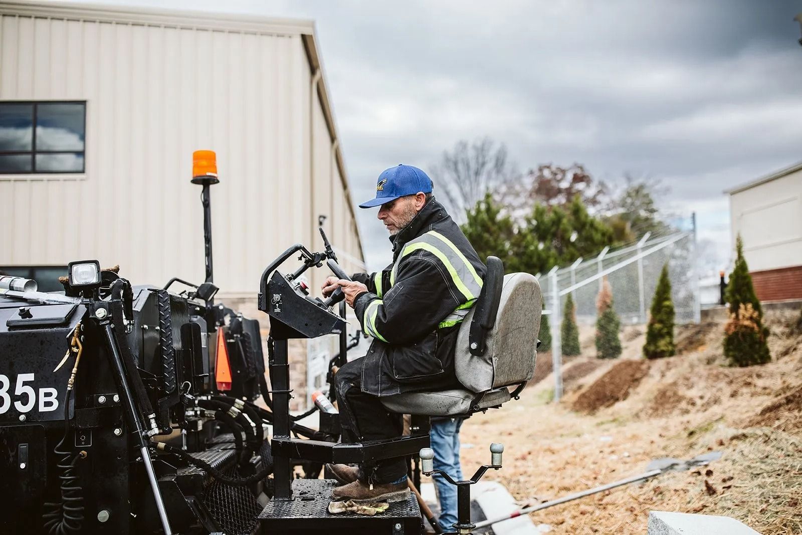 Man operating asphalt paving machine, wearing blue hat, jacket, in front of a building and fence.
