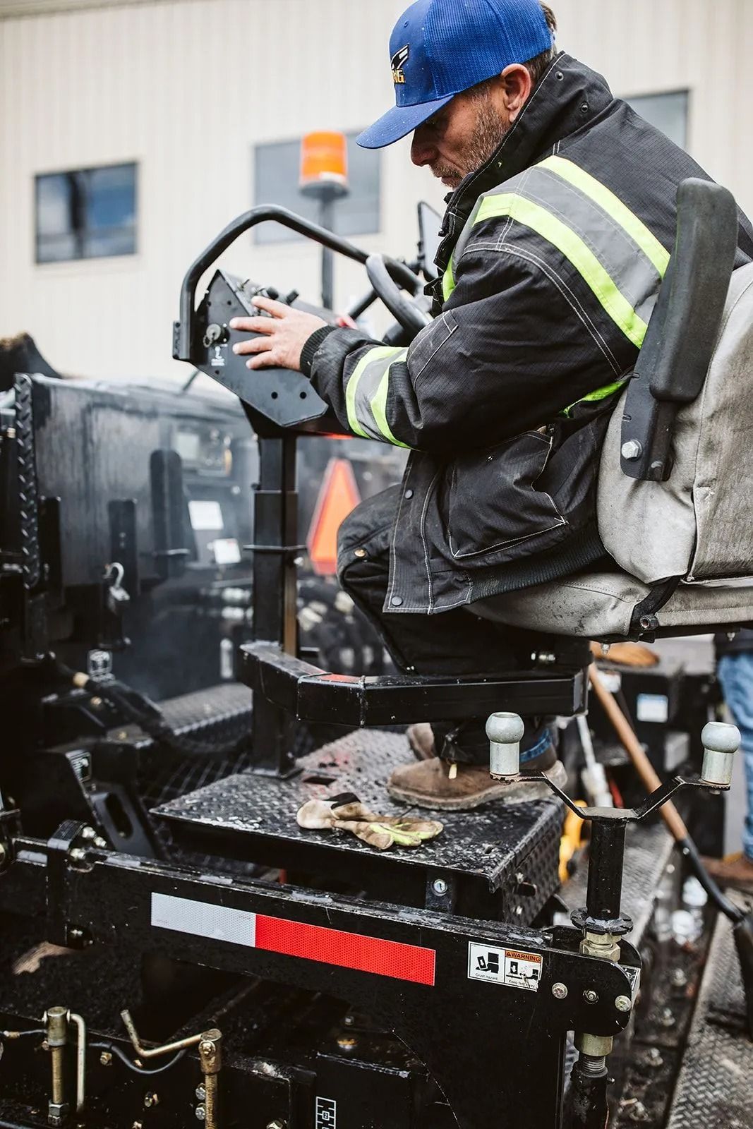Man in blue cap operates heavy machinery outdoors, seated with a dark jacket.