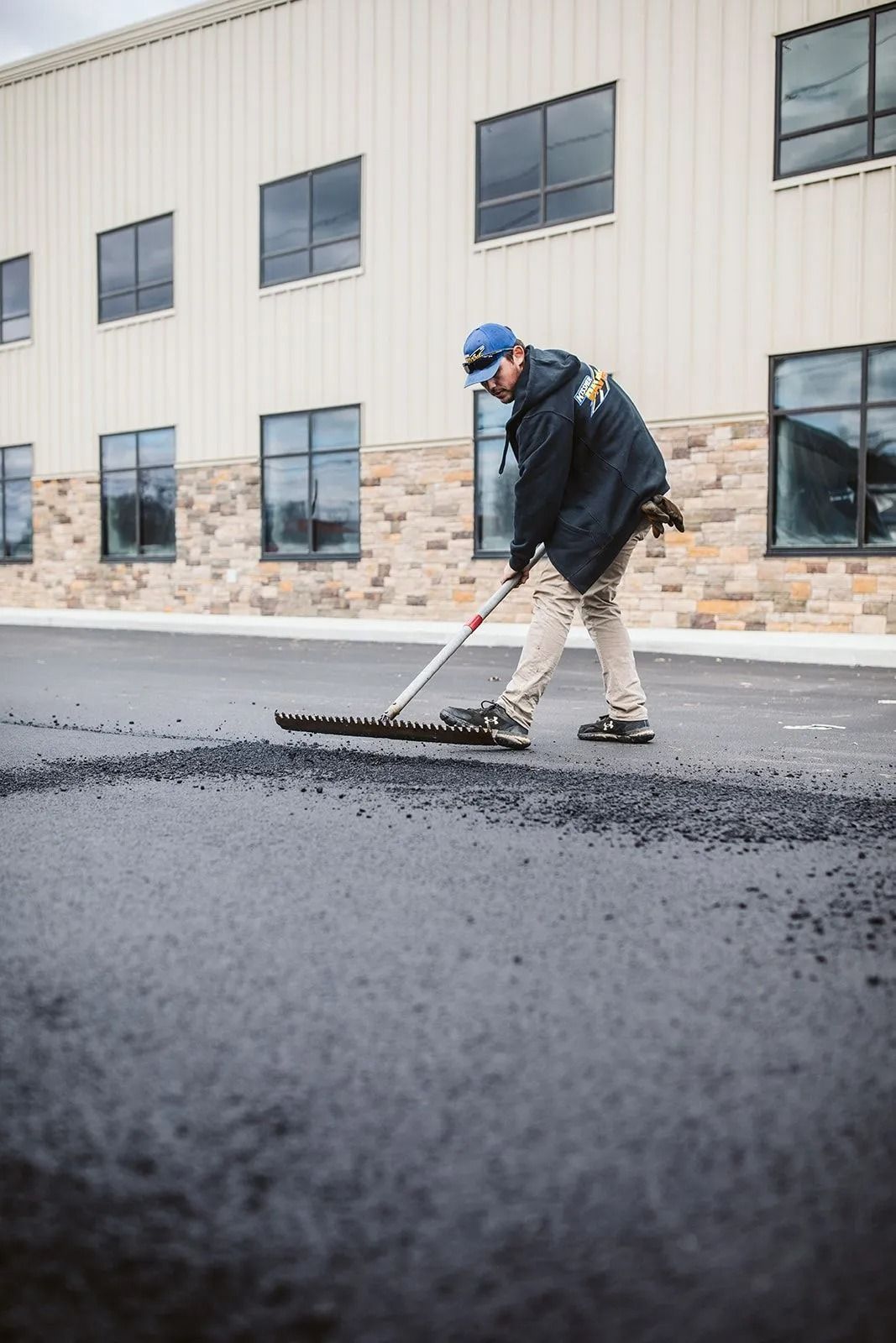 Man raking asphalt on a paved surface in front of a brick building.