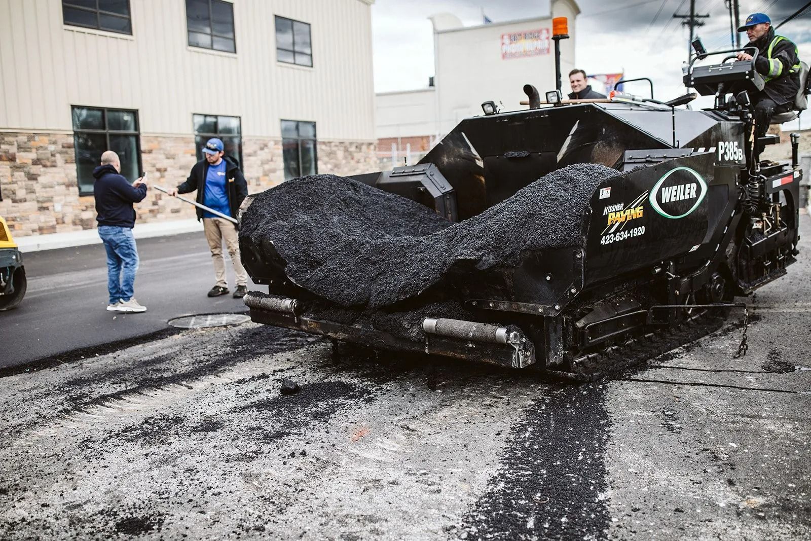 Asphalt paving machine laying new road surface; workers observe.