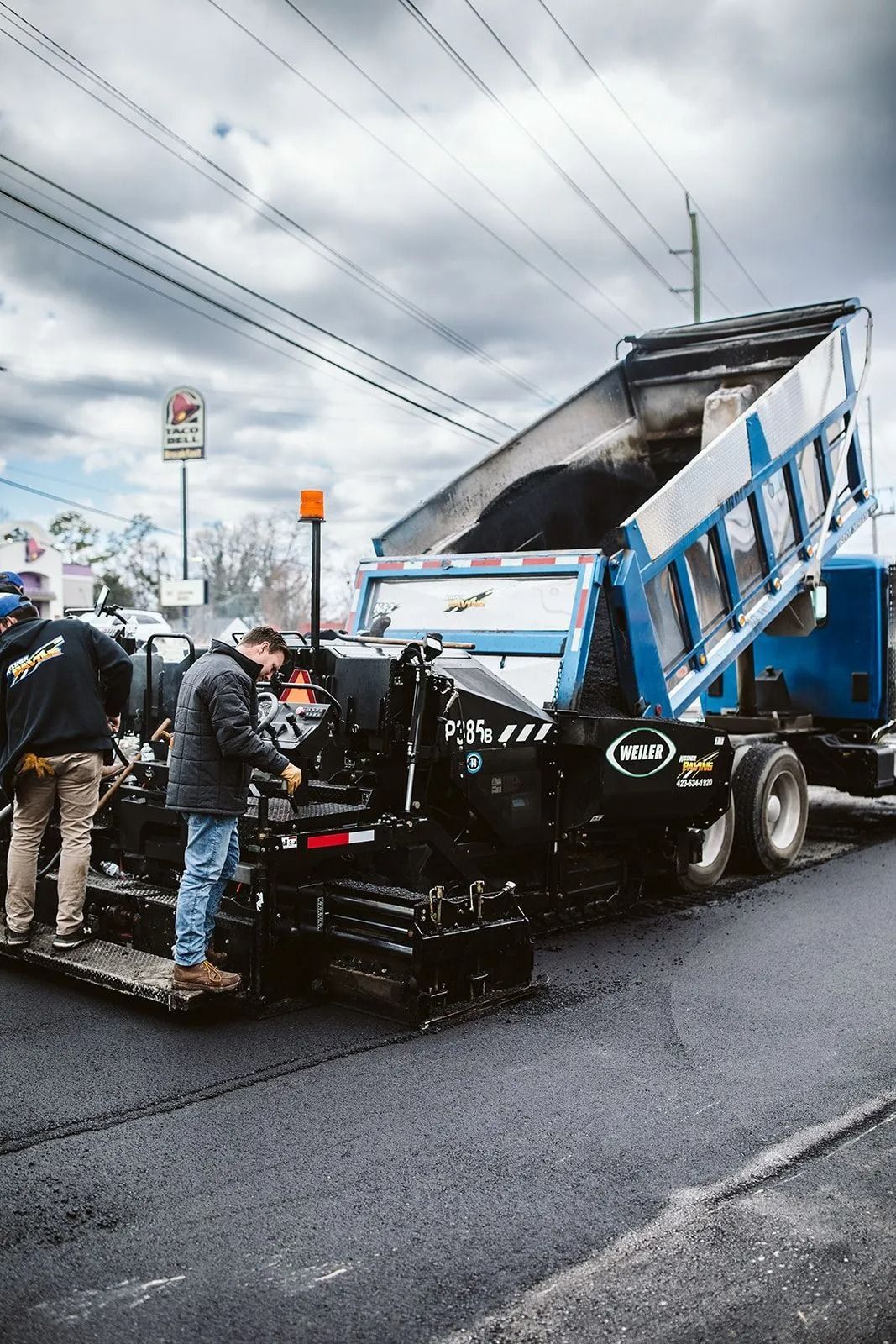 Asphalt paving crew at work; dump truck dumping asphalt into a paver on a road.