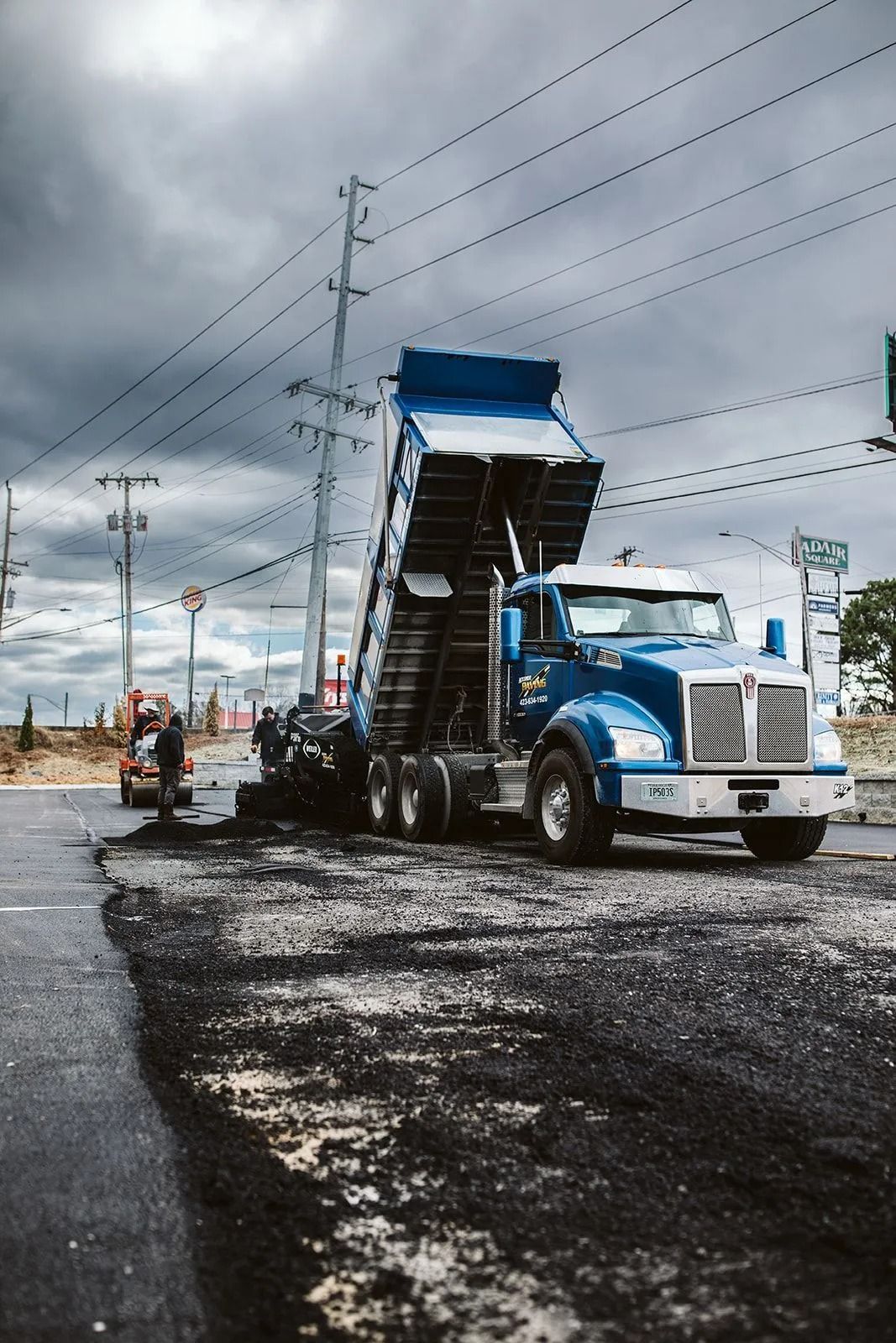 A blue dump truck unloading asphalt onto a road during a road repair under a cloudy sky.