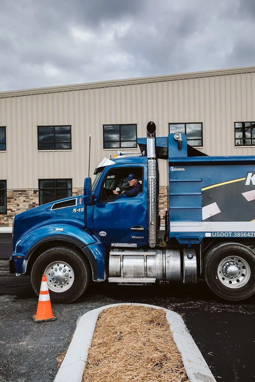 Blue dump truck parked in front of a building with a person driving. A traffic cone and a gravel bed are in view.