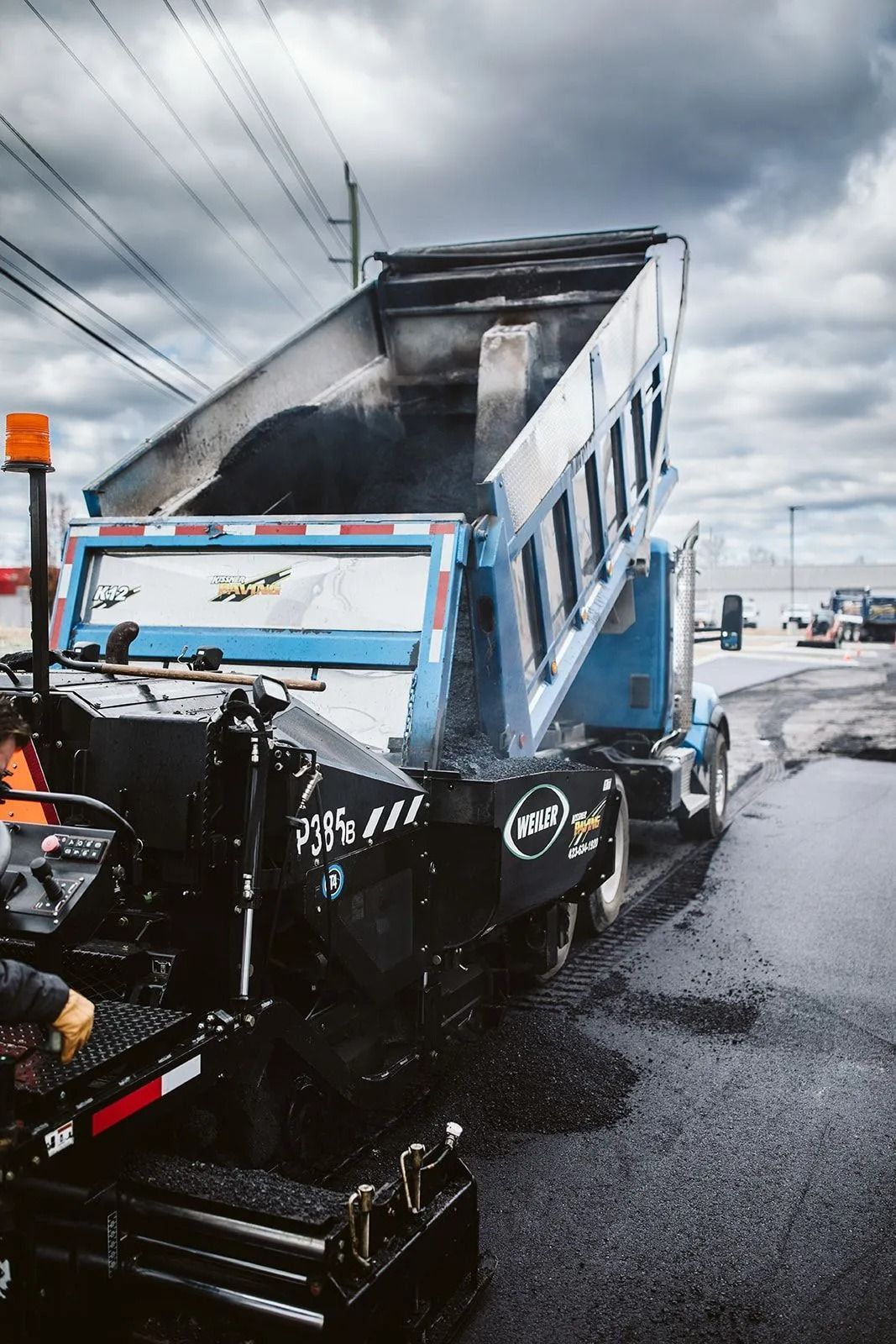 Truck dumping asphalt into a paving machine on a road under overcast skies.