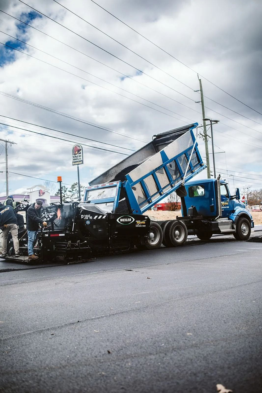 A blue dump truck pouring asphalt onto a road, with workers near a paving machine on a cloudy day.