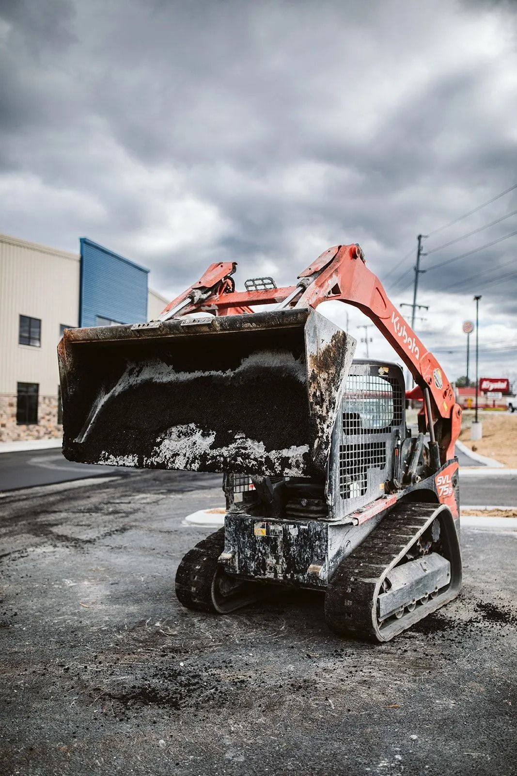 Orange Kubota track loader with a full bucket on a paved area under a cloudy sky.