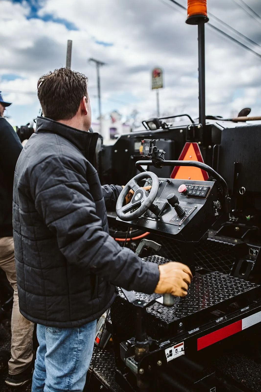 Man operates paving machine, steering wheel and control panel visible. Outdoor setting with cloudy sky.