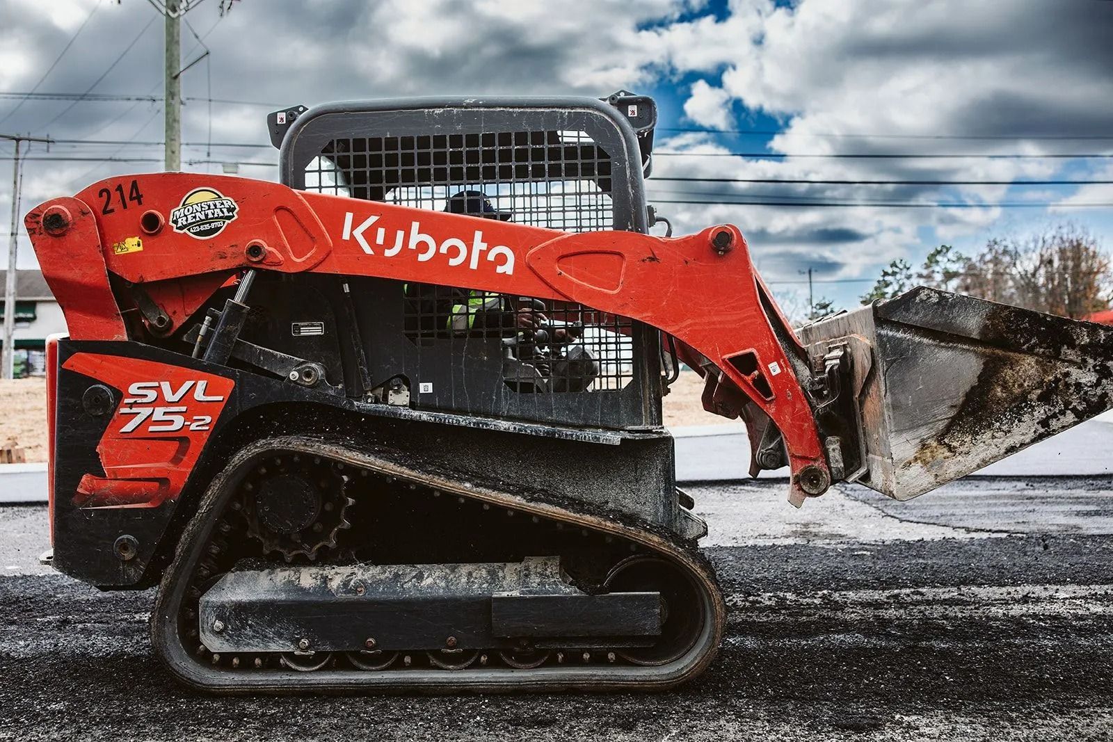 Orange Kubota SVL75-2 compact track loader operating on asphalt, under a cloudy sky.