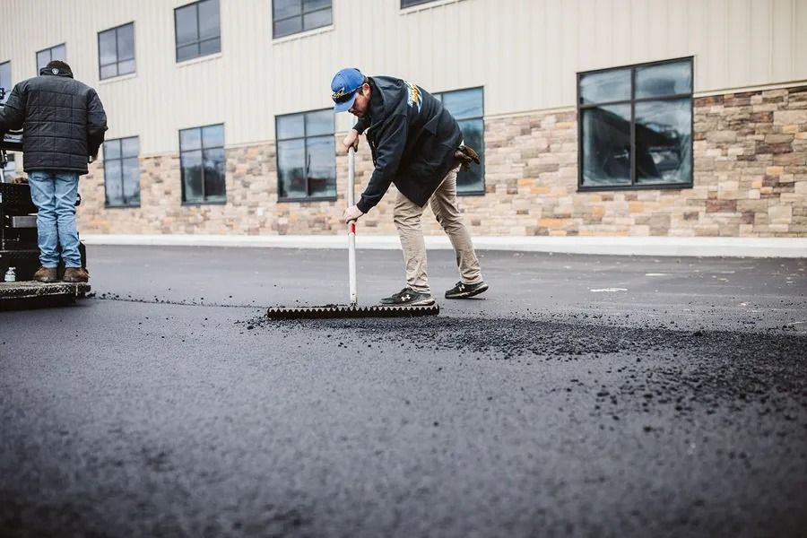 Workers paving asphalt with a large rake in front of a building.