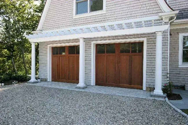 Two wooden garage doors beneath a white-trimmed house exterior with gravel driveway