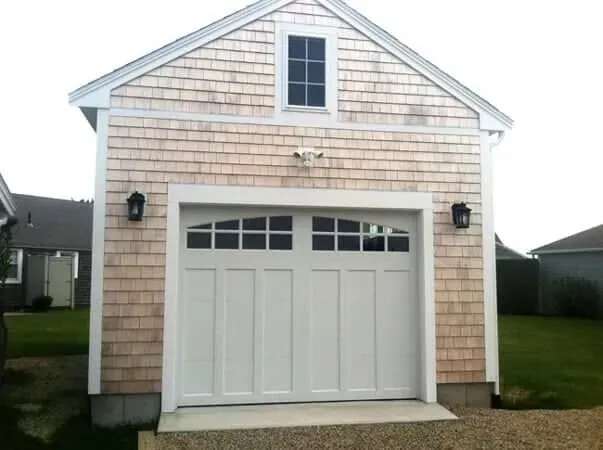White garage with a gabled roof and arched white door on a beige shingled house exterior.