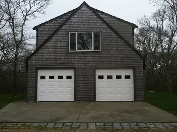 Gray two-car garage with a small window and bare trees behind it