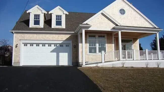 White house with a front porch, garage, and dormer windows beside a driveway under a clear blue sky