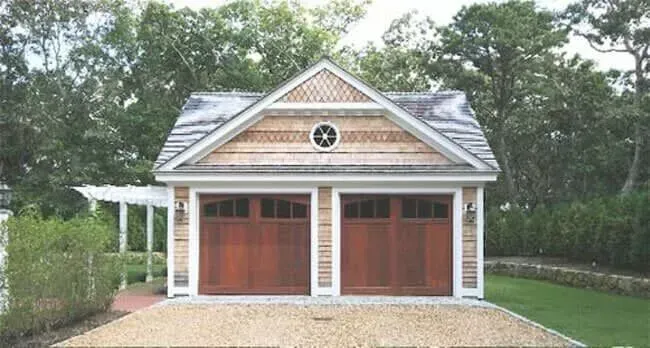 White two-car garage with wooden doors, a circular window, and trees in the background