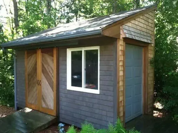Small gray shed with wood-paneled doors and a white window in a wooded yard