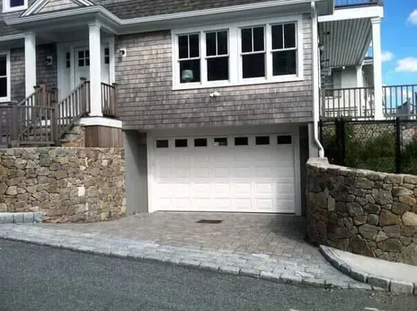 Stone house front with white garage door under a raised driveway, flanked by stone retaining walls.