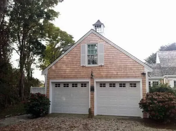 Two-car garage with beige siding, white doors, and a small cupola, surrounded by trees and shrubs.