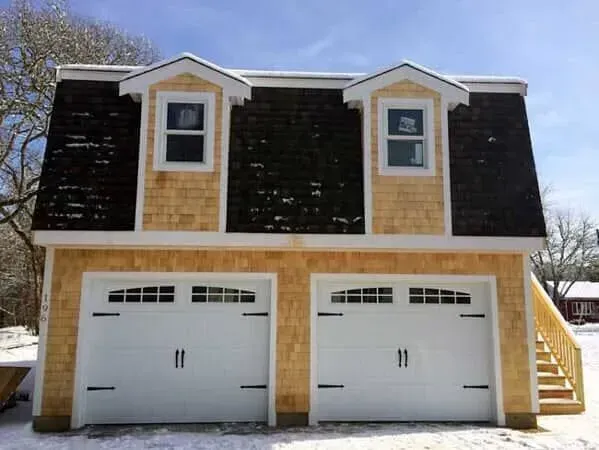 Two-car garage with tan siding, black roof, and two dormer windows in a snowy yard
